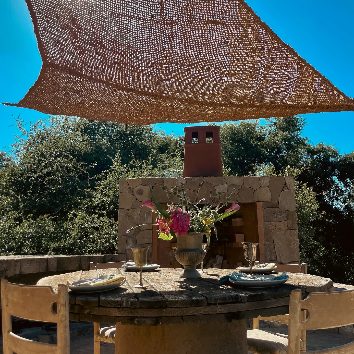 Elongated triangle coconut fiber shade sail above an outdoor stone dining area with flowers and rustic furniture.