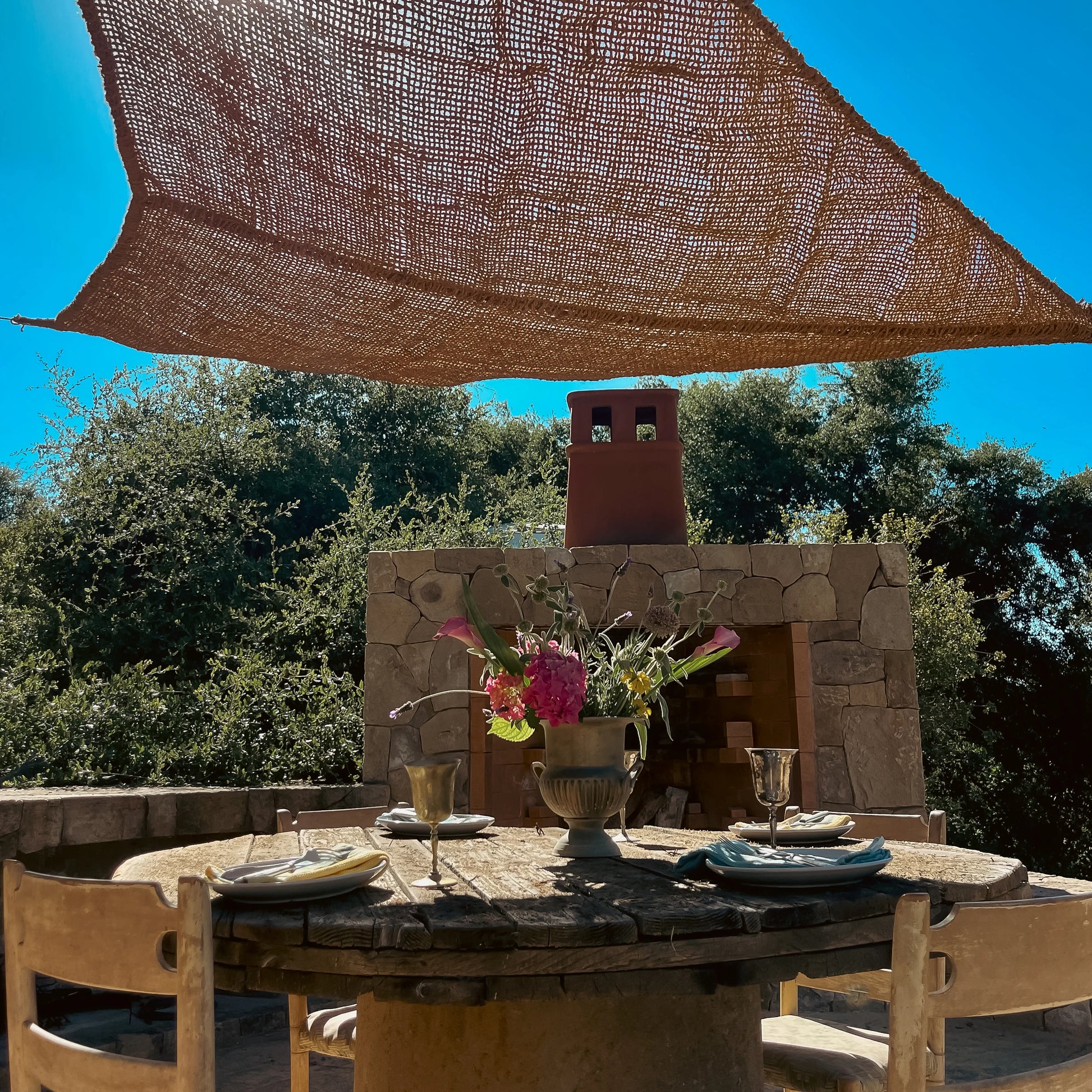 Elongated triangle coconut fiber shade sail above an outdoor stone dining area with flowers and rustic furniture.