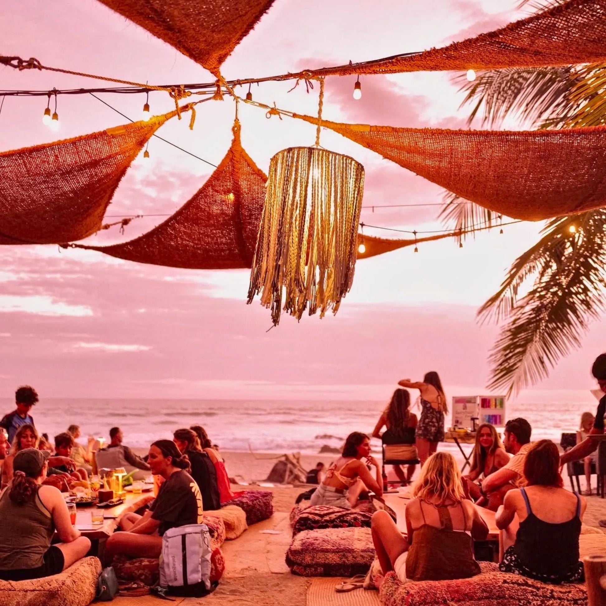 People enjoying a sunset at a beachside gathering with palm trees and string lights. 12' x 12' x 12' triangle coconut fibre shade sail natural fibers handmade artisan europe rustic vibes non toxic rooftop bar  coco du sol beach party boho chic