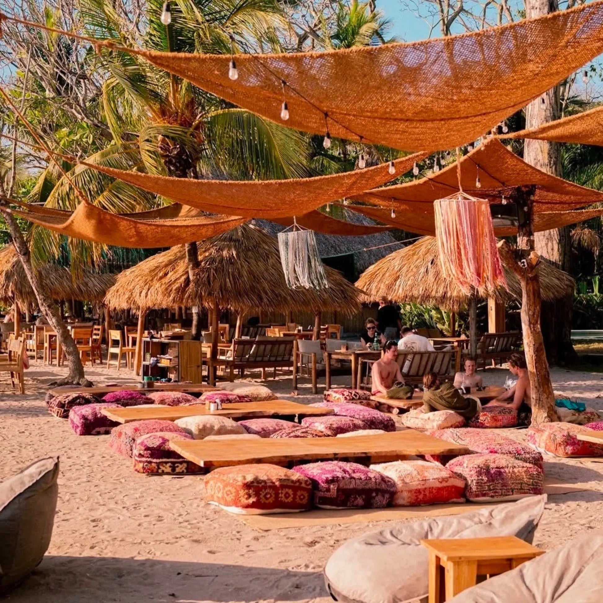 Beachside setting with thatched umbrellas, lounge chairs, and people enjoying a sunny day. 12' x 12' x 12' triangle coconut fibre shade sail natural fibers handmade artisan beach rustic vibes non toxic costa rica coco du sol chic living dappled light breathable 