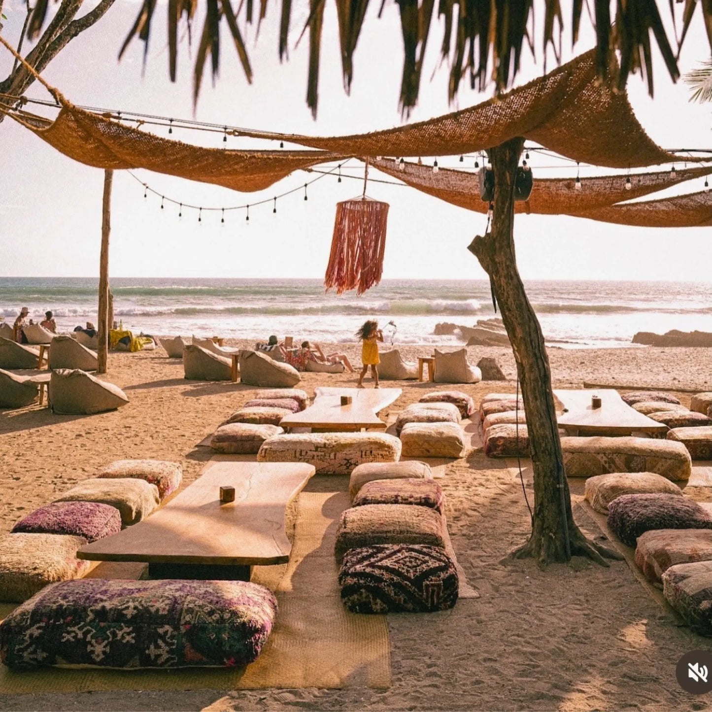 Beachside seating area with tables and cushions under a thatched roof, with people in the background. 12' x 12' x 12' triangle coconut fibre shade sail natural fibers handmade artisan europe rustic vibes non toxic coco du sol filtered dappled light breathable shade pattens costa rica