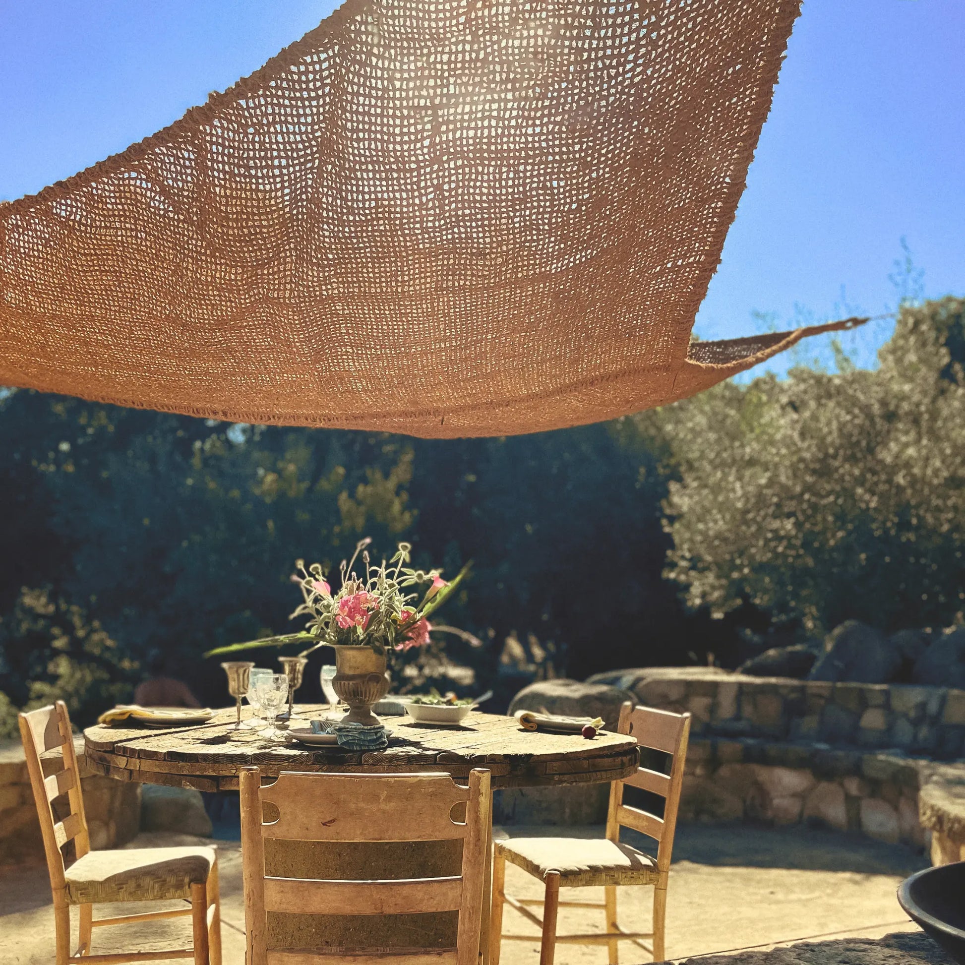 Outdoor dining setup with a round table and chairs under a large woven canopy. 12' x 12' x 12' triangle coconut fibre shade sail natural fibers handmade artisan europe rustic vibes non toxic light filtered coco du sol chic living dappled light breathable 
