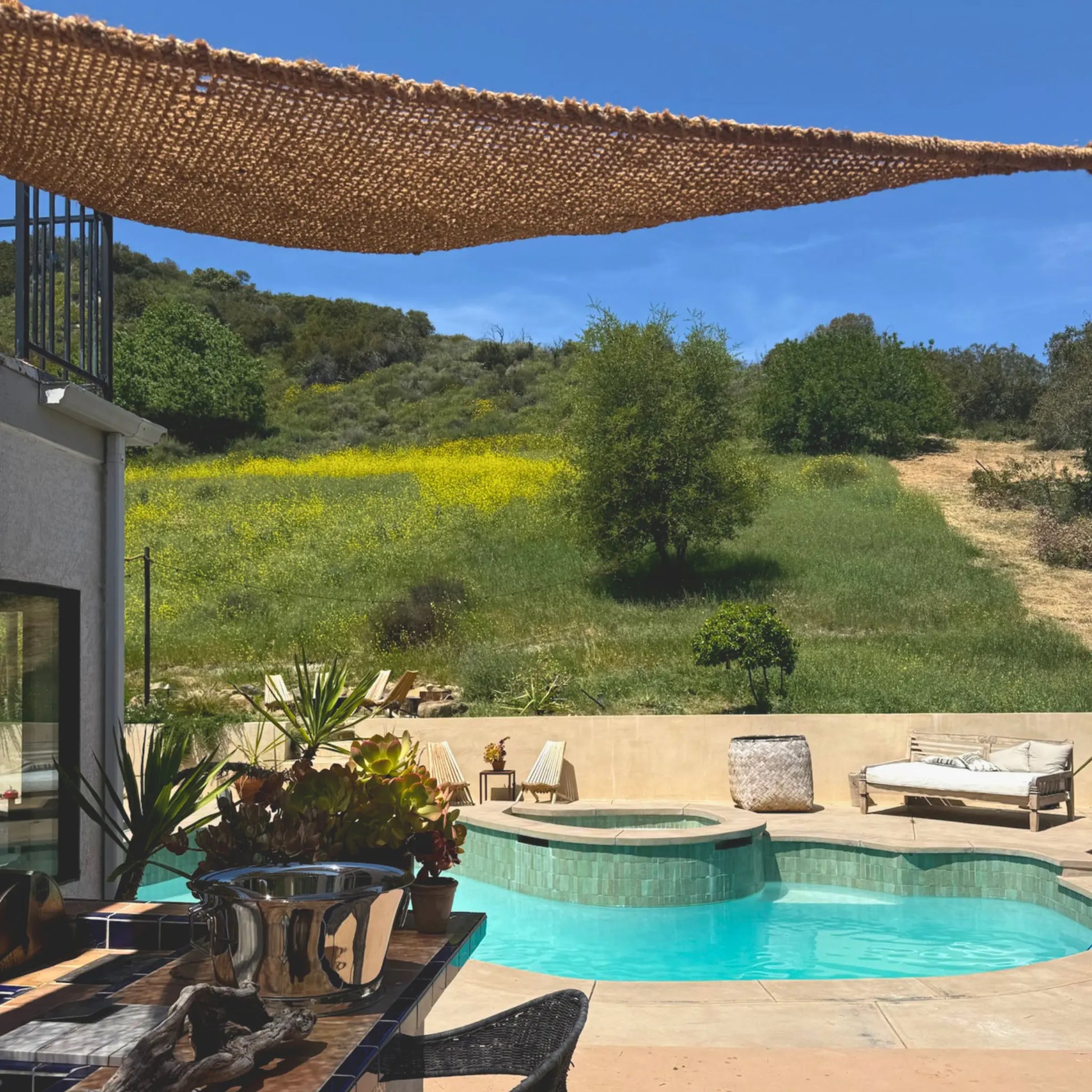 Outdoor pool area with lounge chairs, plants, and a view of greenery. 12' x 12' x 12' triangle coconut fibre shade sail natural fibers handmade artisan europe rustic vibes non toxic rooftop bar topanga california coco du sol filtered dappled light breathable shade pattens 